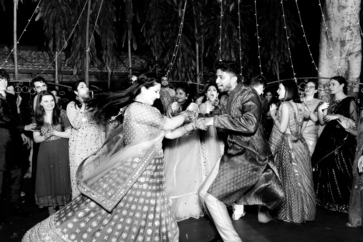 In black and white, a woman spins in a swirling lehenga beside a man dancing in a dark kurta