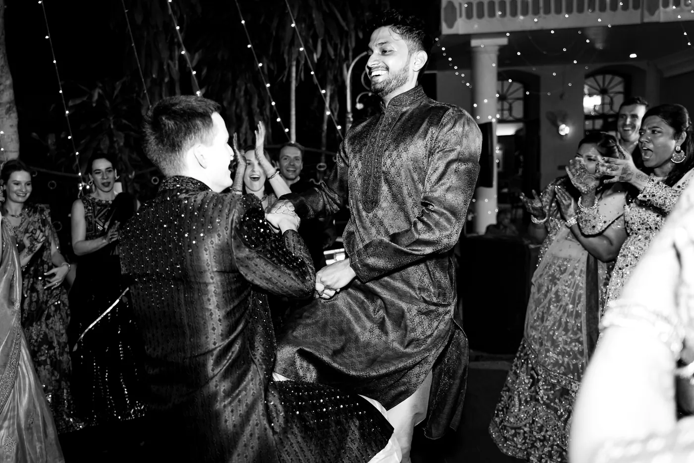 In black and white, a man laughs with one leg lifted mid dance as friends move around him
