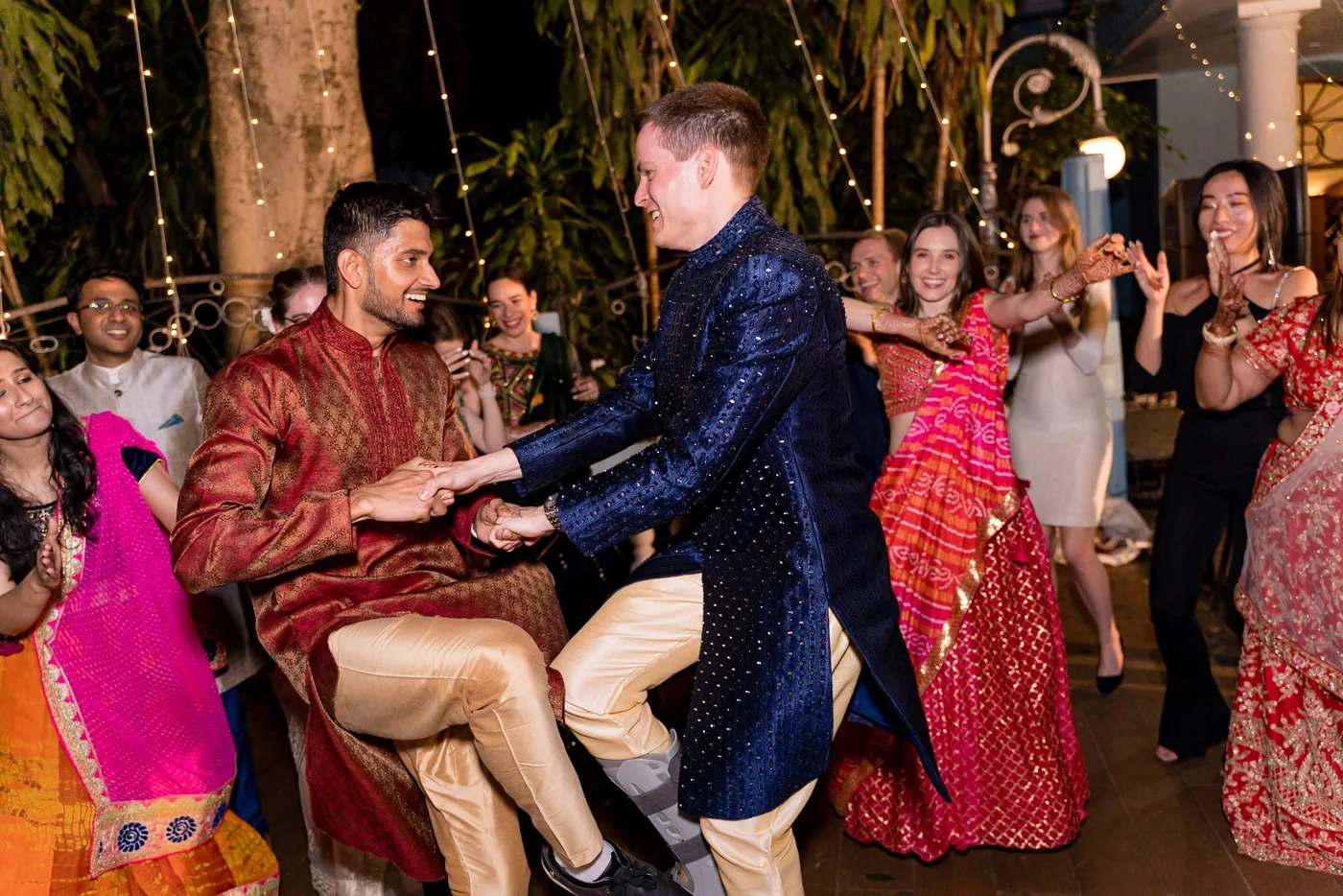 Stefan in navy dances with a man in a red sherwani, hands joined, as women in pink dance behind