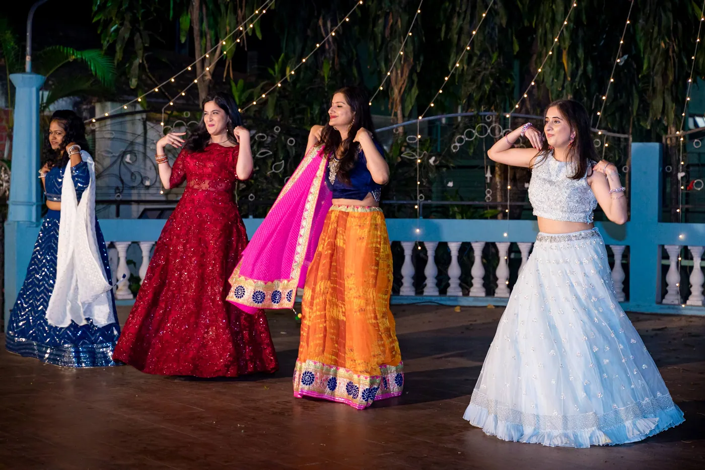 Four women in red, orange and pale blue lehengas dance across the dark wooden floor
