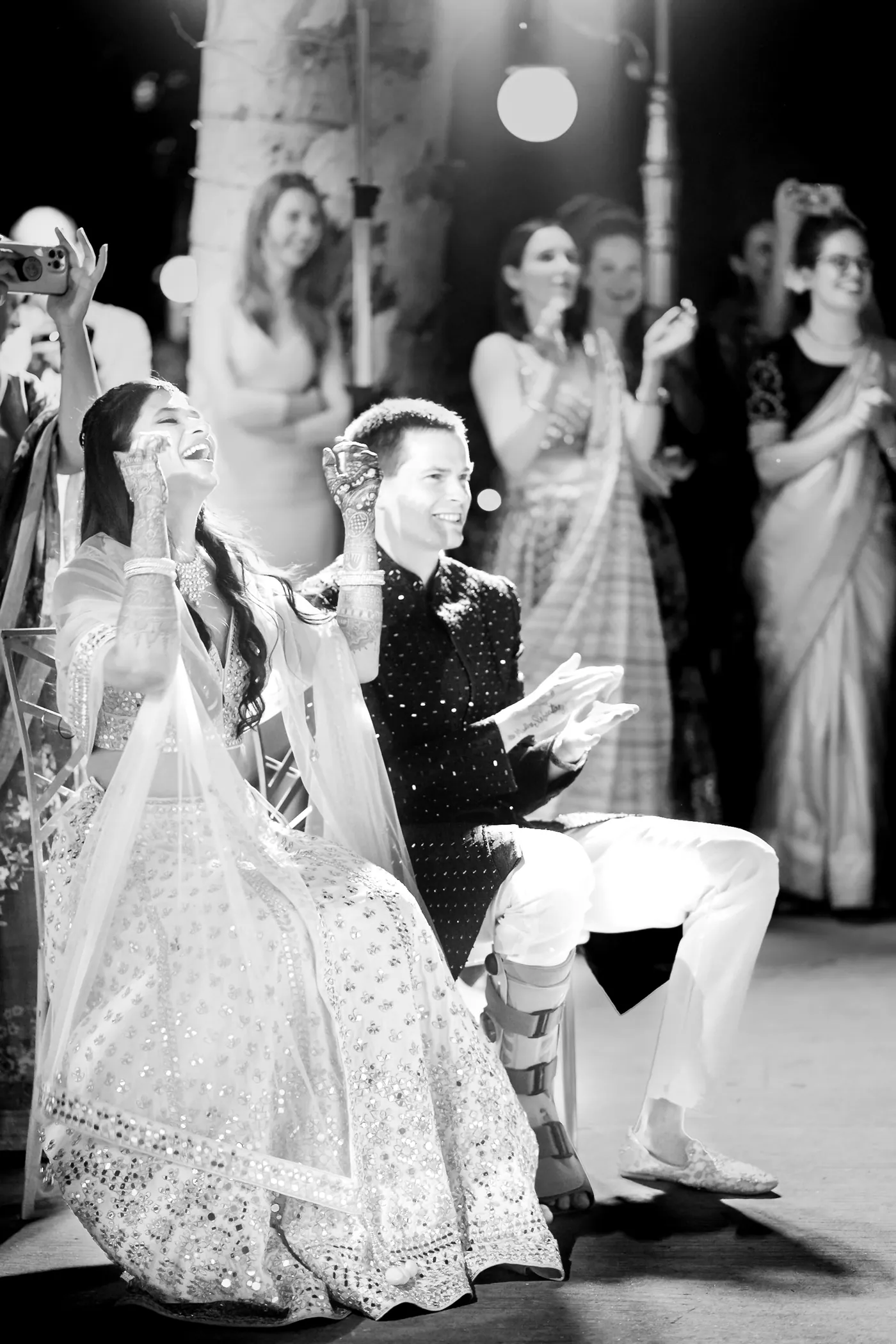 In black and white, a woman in a full lehenga twirls while a man in a suit sits close by