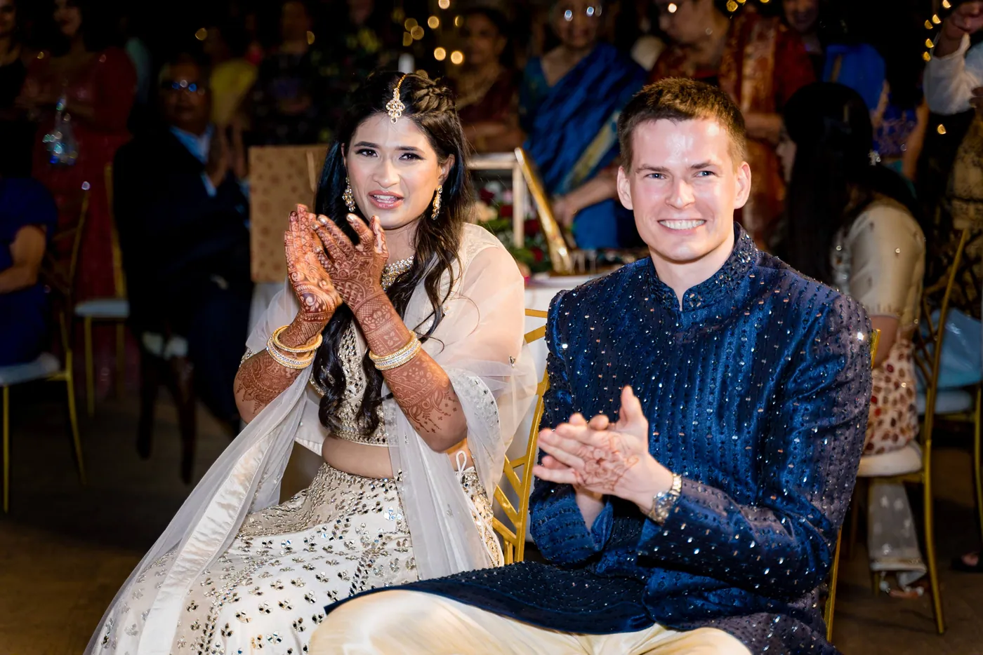 Medha with henna on her hands and Stefan in navy clap along from their seats during the performances