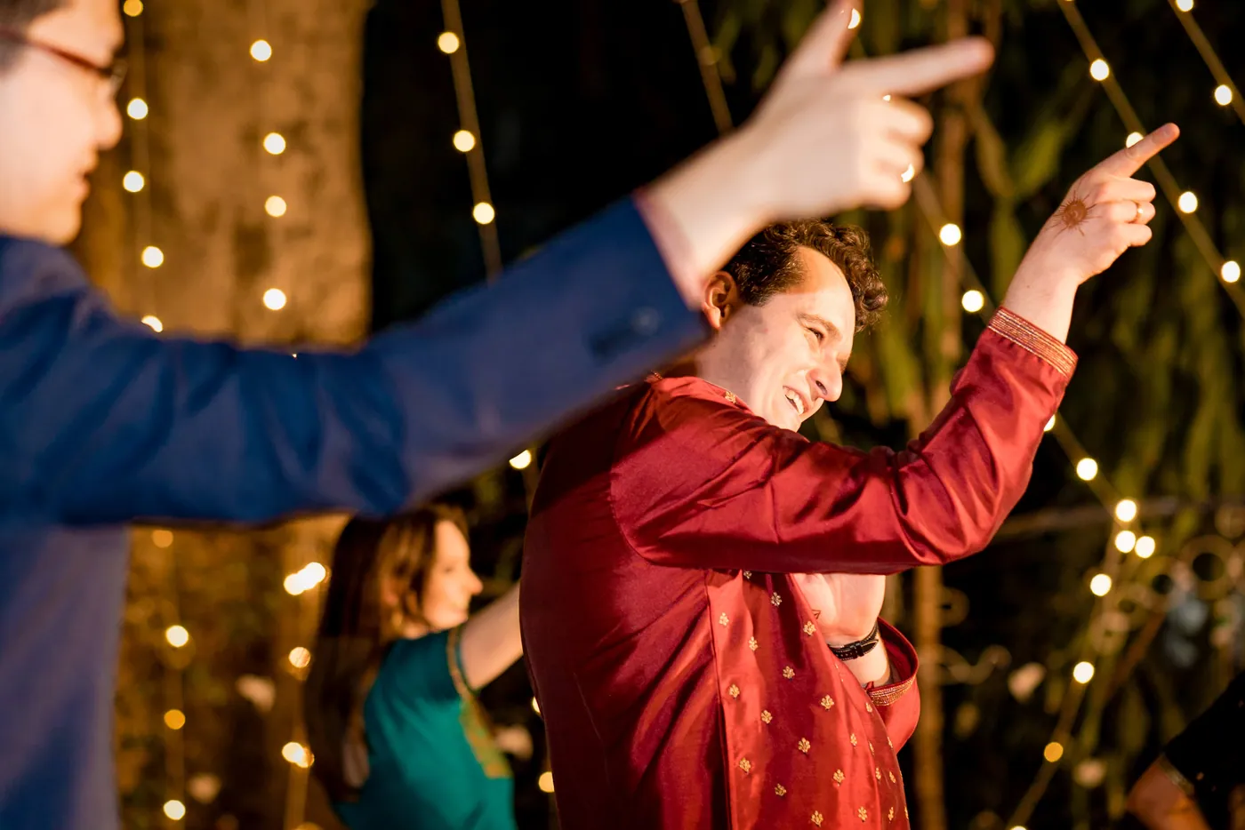 A man in a red kurta points with both arms outstretched while another guest points back