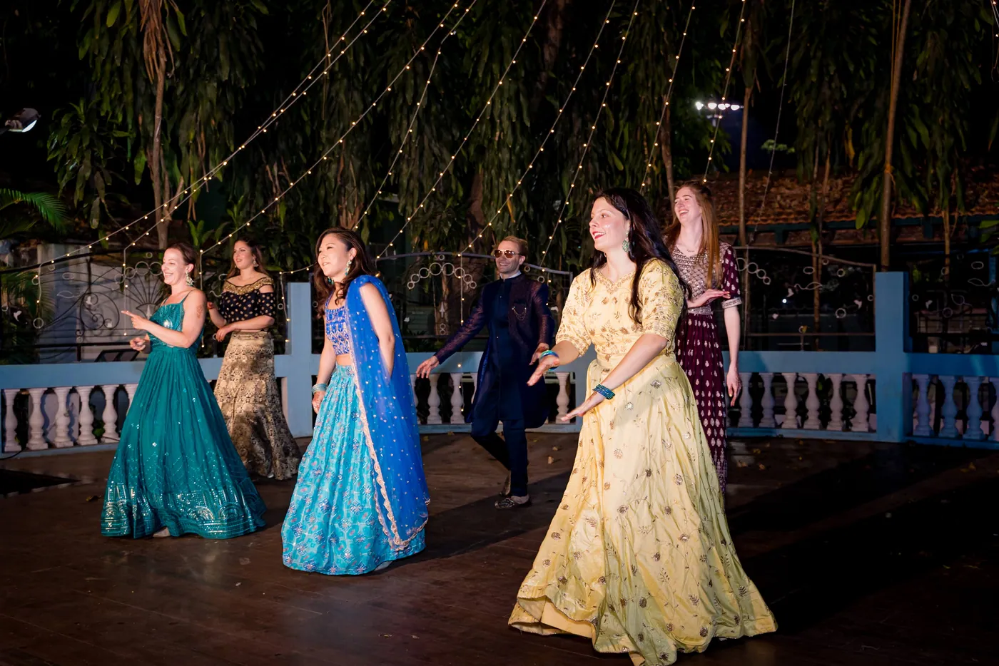 Women in teal, blue and yellow lehengas dance side by side with a man in sunglasses behind them