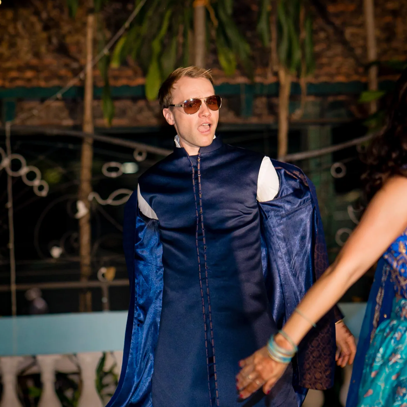 A man in a navy sherwani and sunglasses mouths the lyrics mid dance