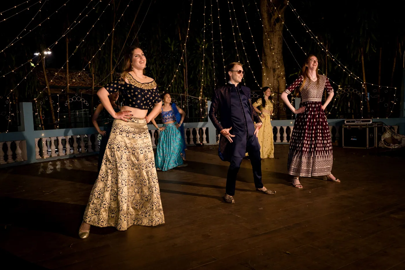 Dancers in lehengas and a man in dark sunglasses pose with hands on hips on the dark floor
