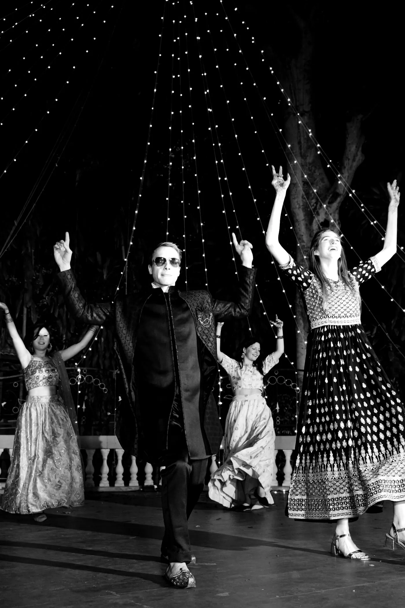 In black and white, a man in sunglasses strikes a pose and two women dance beside him