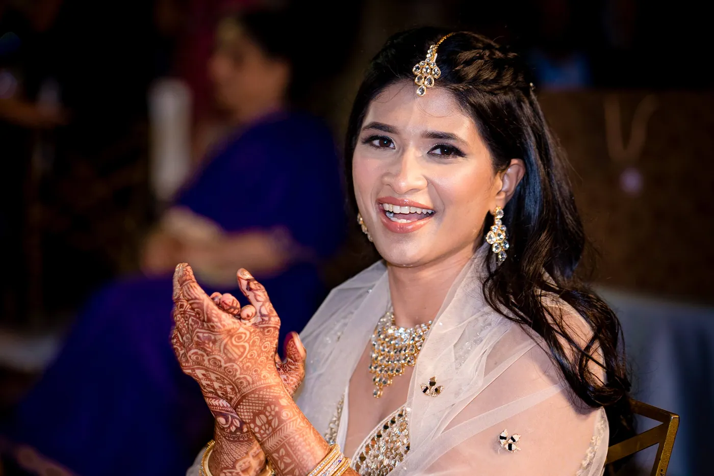 Medha smiles to the camera with hands raised in a clap, her mehendi bright in the frame