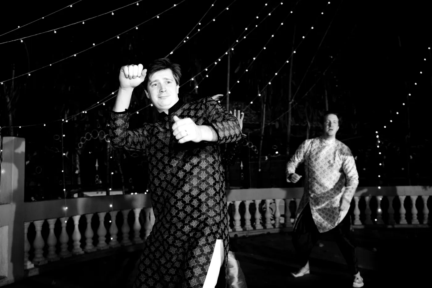 In black and white, a man in a patterned jacket dances hard with a raised fist