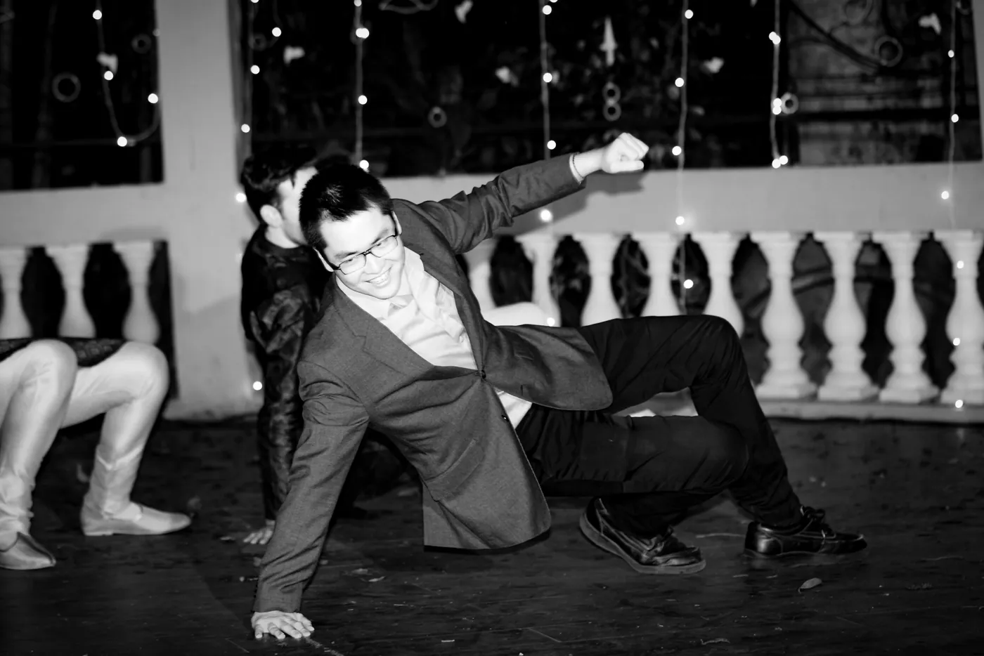 In black and white, a man in a grey jacket drops to the floor mid dance as others crouch