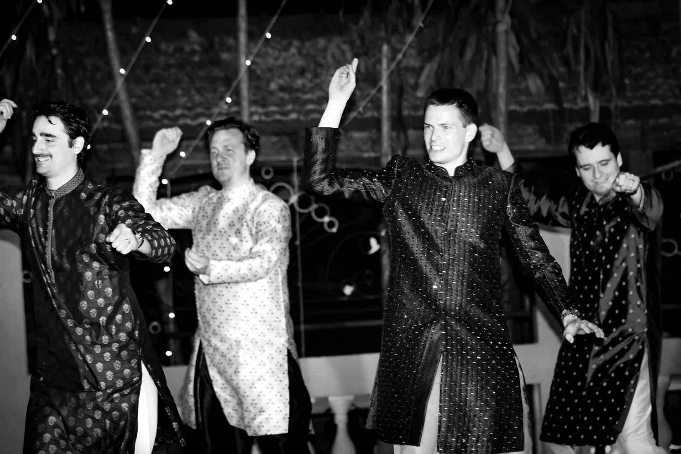 In black and white, a row of men in patterned sherwanis dance in unison