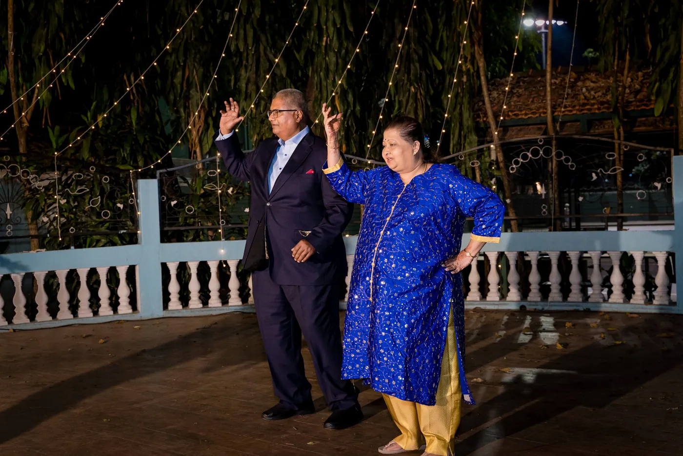 A man in a dark suit and a woman in a blue kurta dance together beside the blue railing