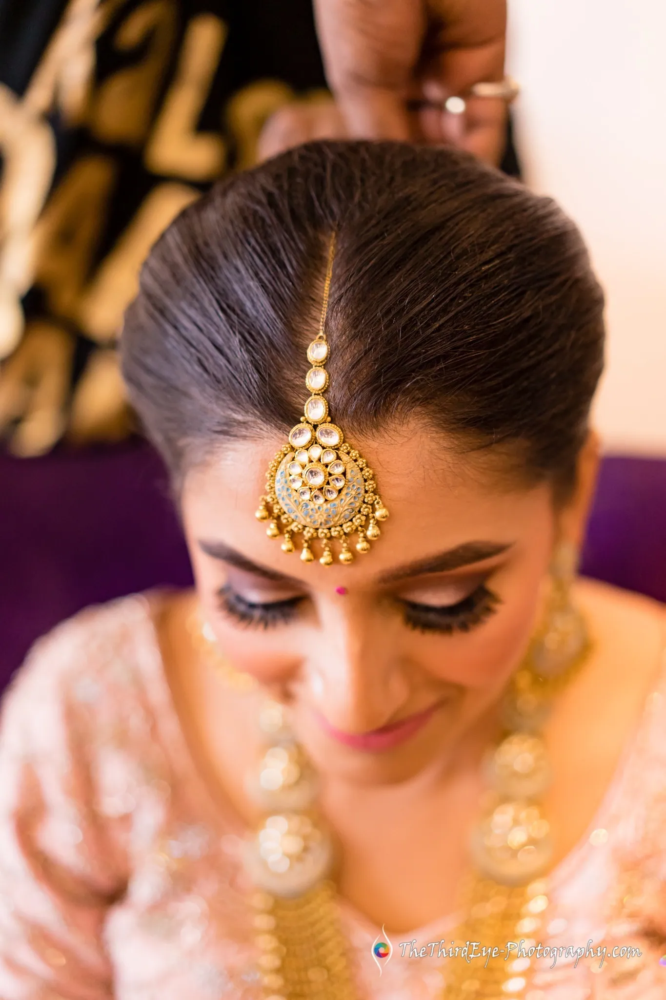 The bride looks down as a hand adjusts her gold maang tikka, layered necklaces glowing in warm light