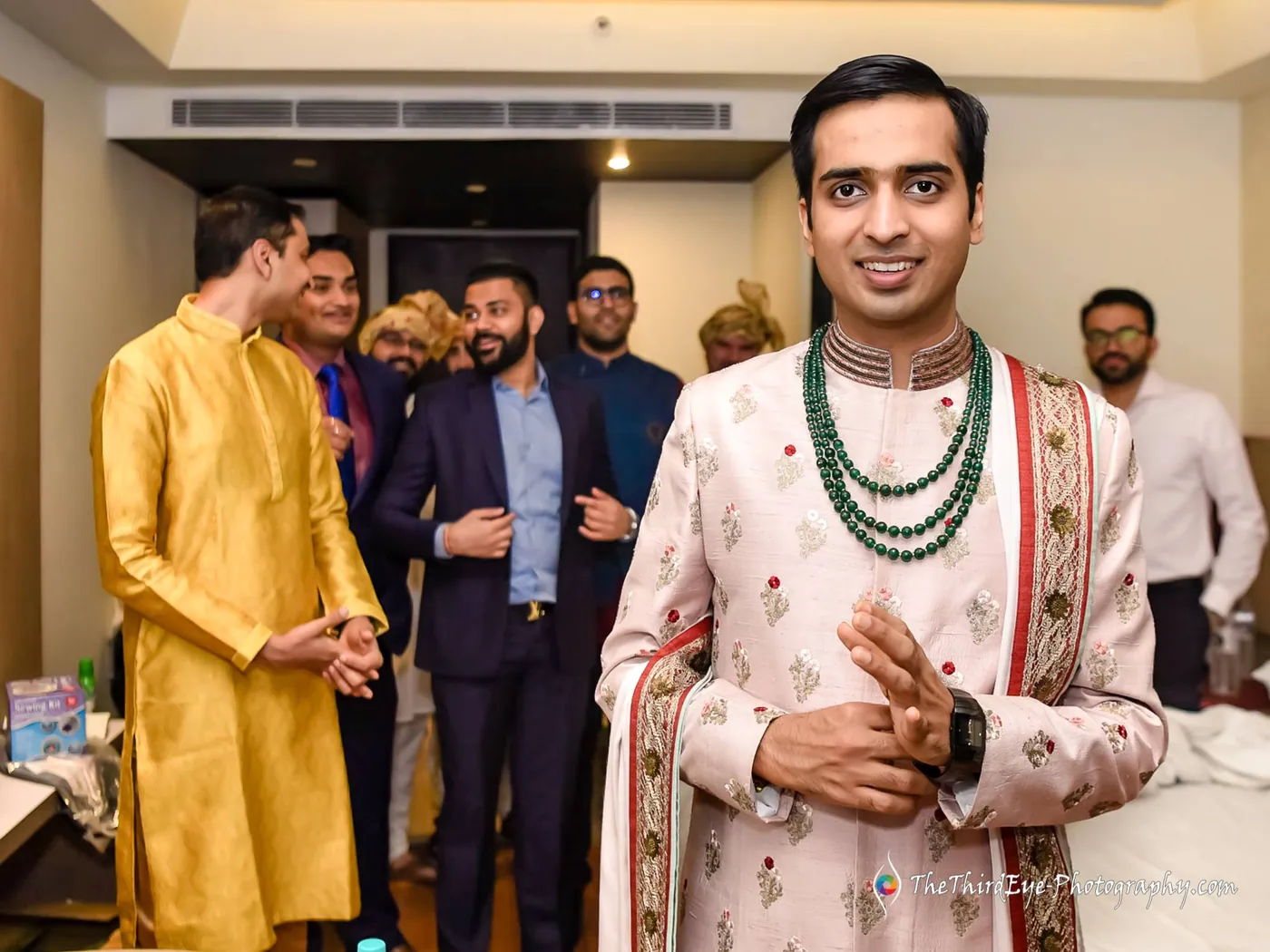 The groom in a pale embroidered sherwani with green beads stands smiling while friends gather behind him