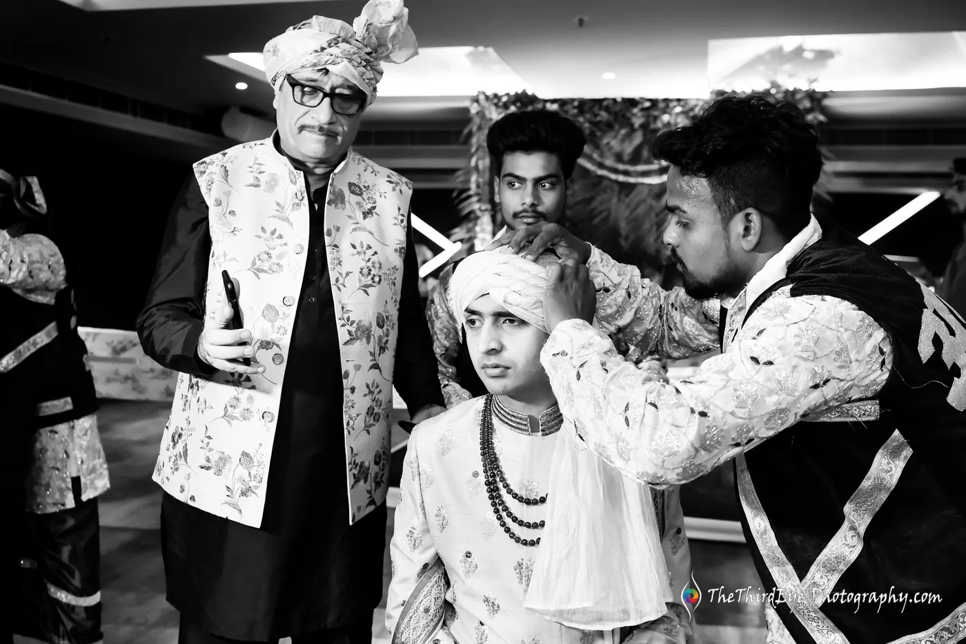 Black and white men help tie a turban on the groom as an older man in a patterned jacket watches