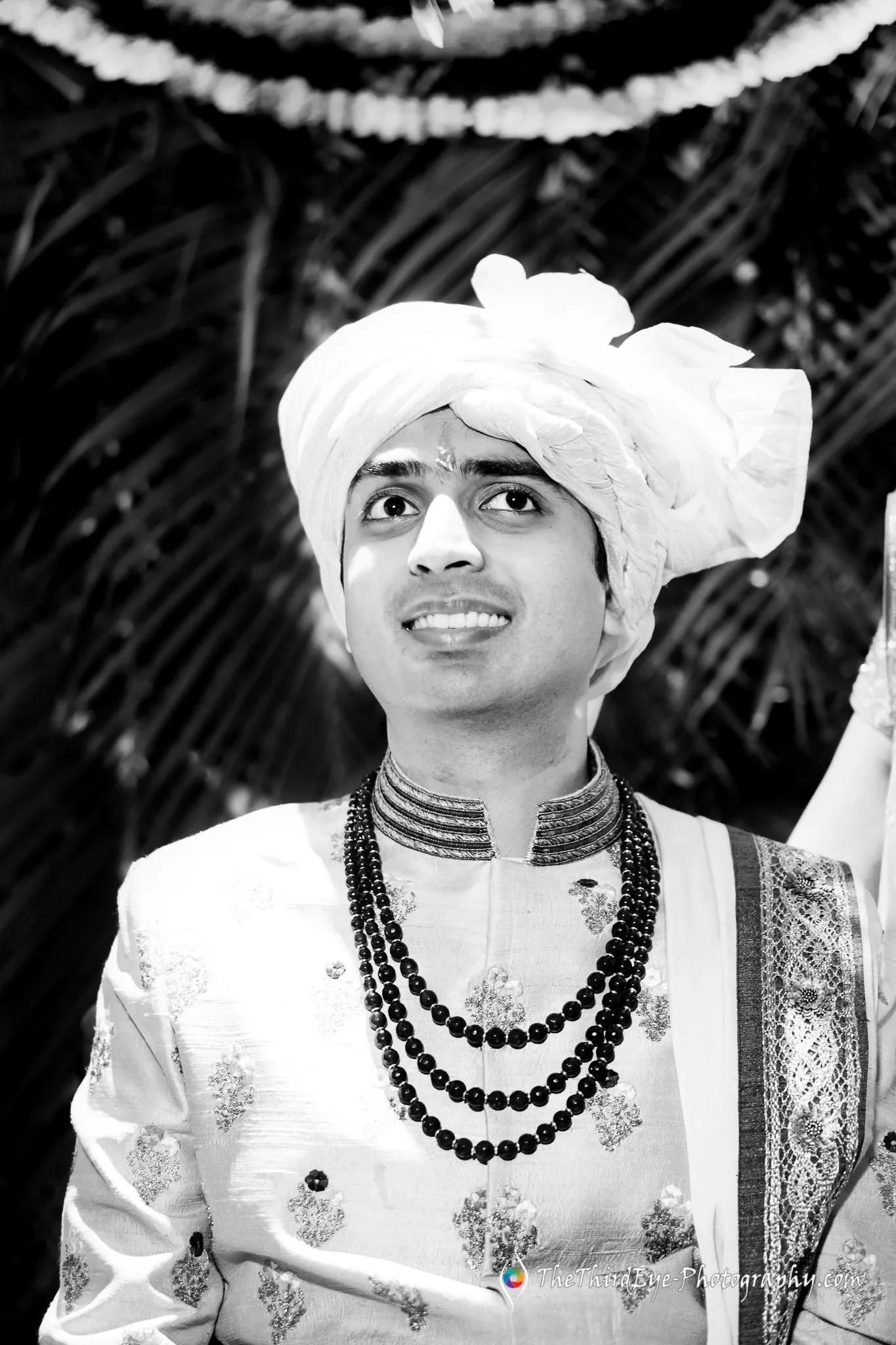 Black and white portrait of the groom in a white turban and layered beaded necklaces, looking up and smiling