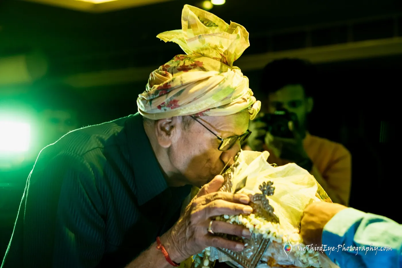 An older man in glasses presses his forehead to a yellow turbaned head in a blessing, lit by green light
