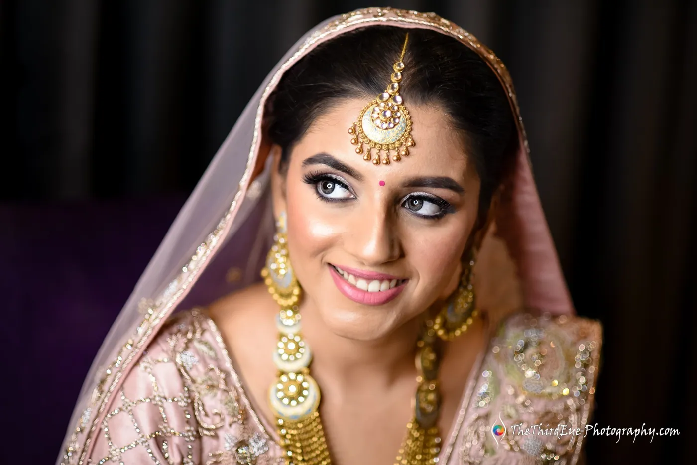 The bride in a pale pink lehenga and gold jewellery looks sideways with a bright smile