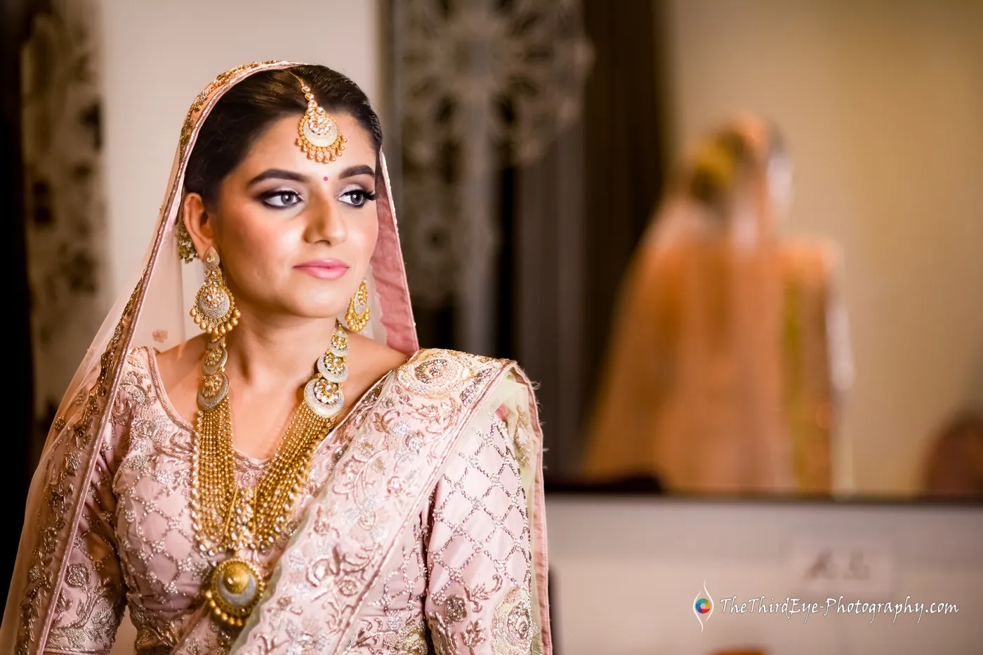 The bride sits in her pale pink bridal outfit, gold necklaces draped, an orange glow softening the background