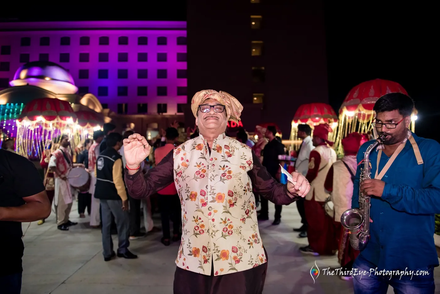 An older man in a pastel kurta dances with arms out at night, decorated horse canopies glowing behind him