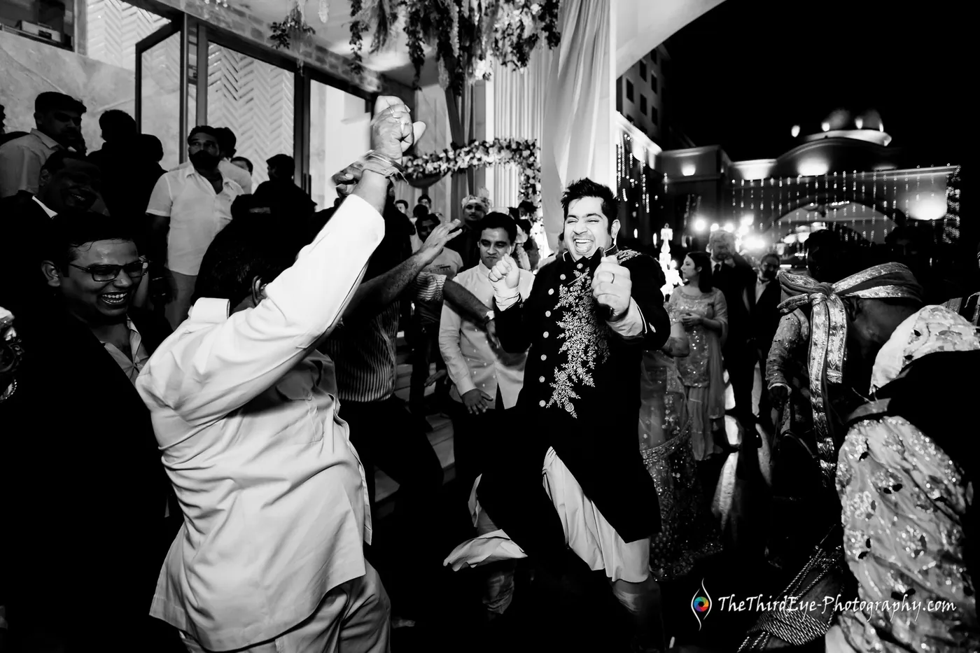 Black and white crowd dancing outside at night, a man in a dark jacket mid step at the centre