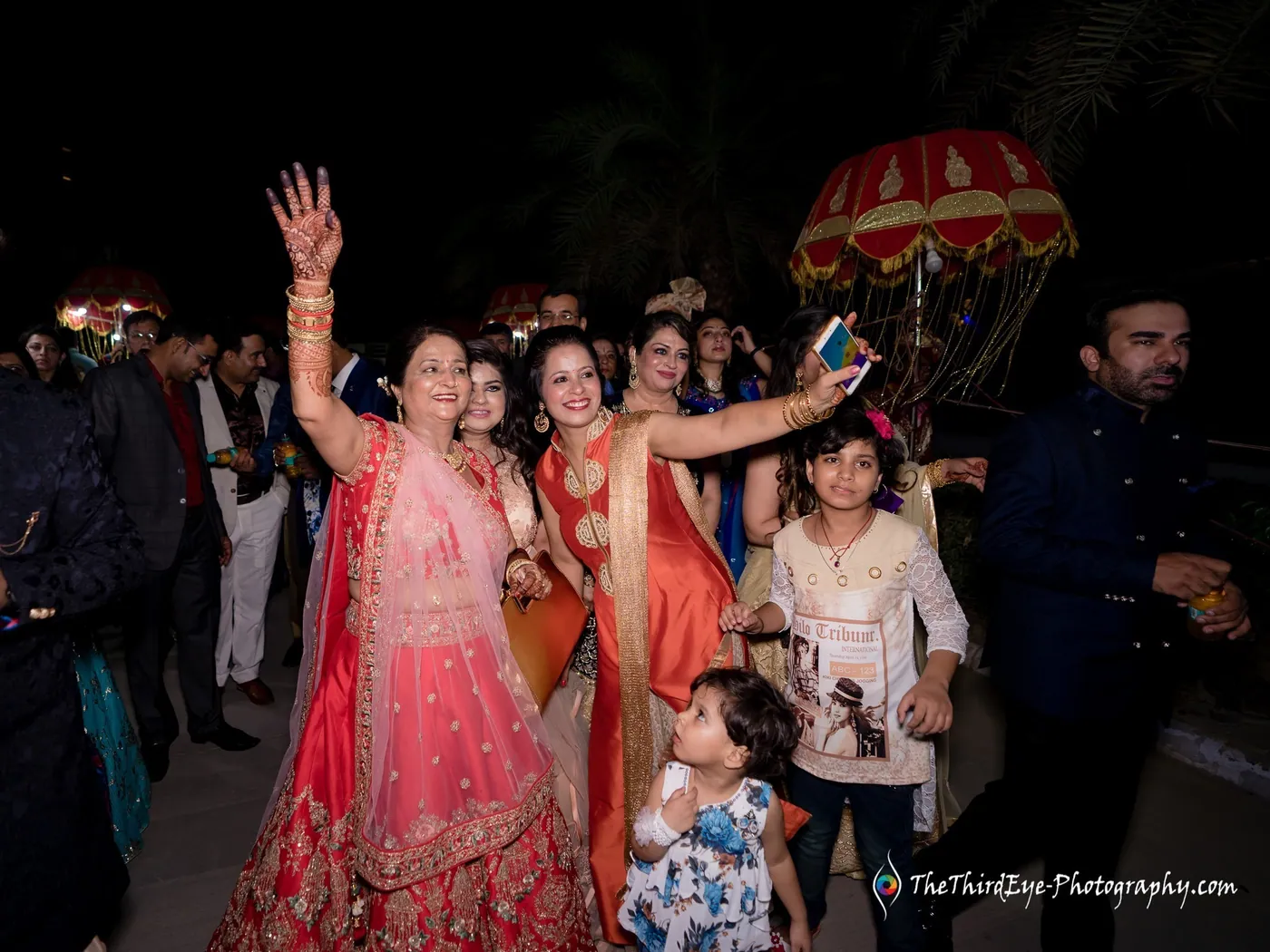 Women in bright lehengas cheer with arms raised, one taking a selfie, as a small child stands in front