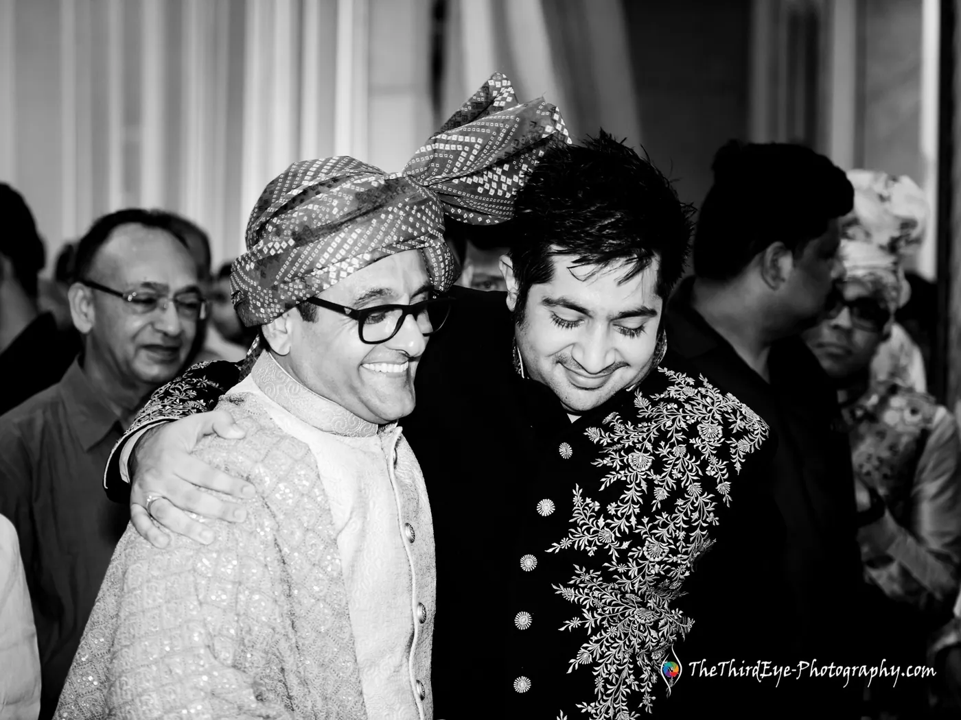 Black and white frame of two men in turbans embracing, one beaming, in the middle of a crowd