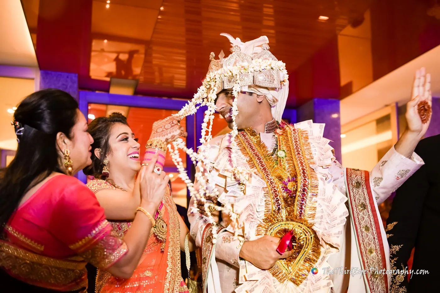 The groom in a white sehra and gold necklaces is welcomed by a laughing woman as a man waves
