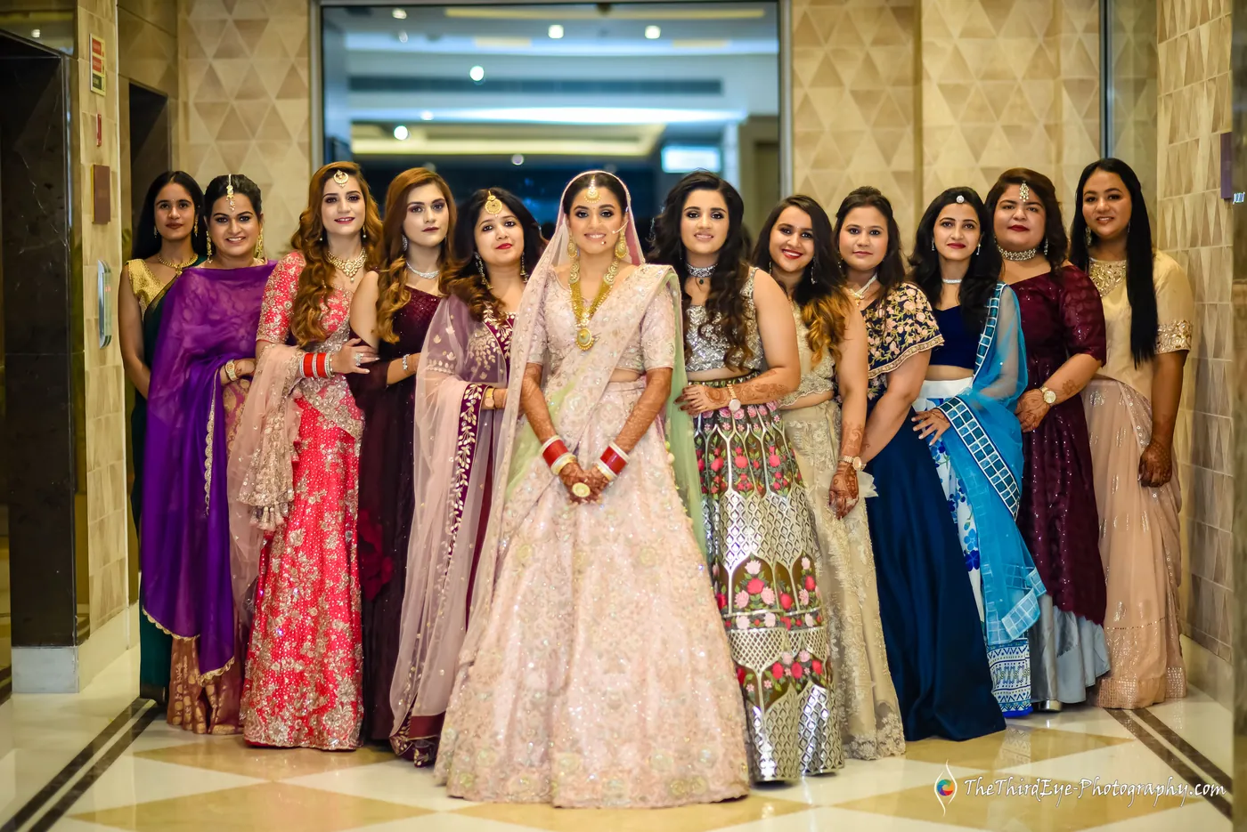 The bride stands among women in bright outfits lined up in a hotel lobby, all facing the camera