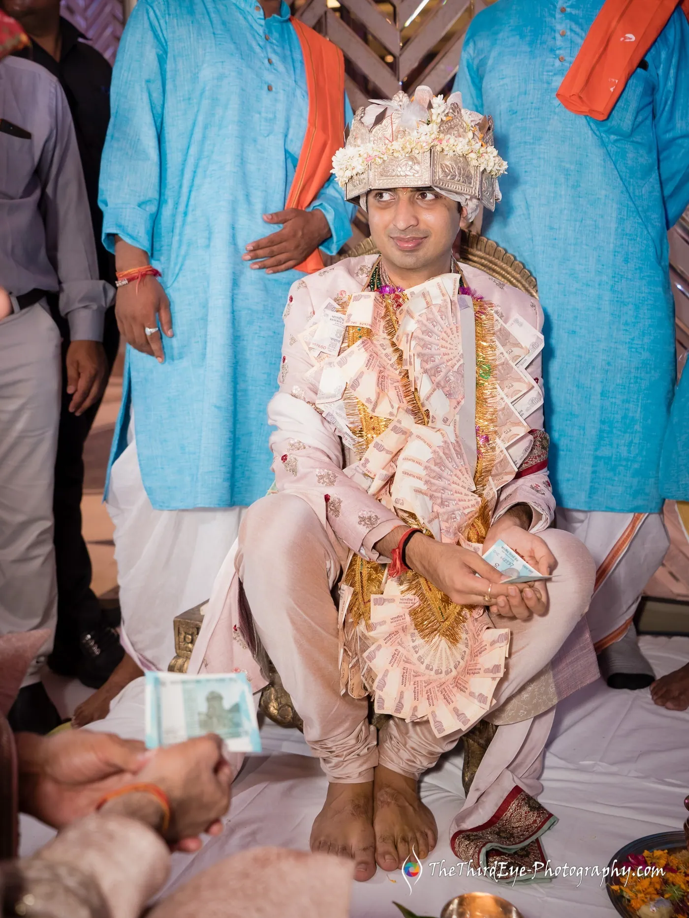 The groom sits cross legged in white and gold, men in blue and orange standing around, notes on the floor