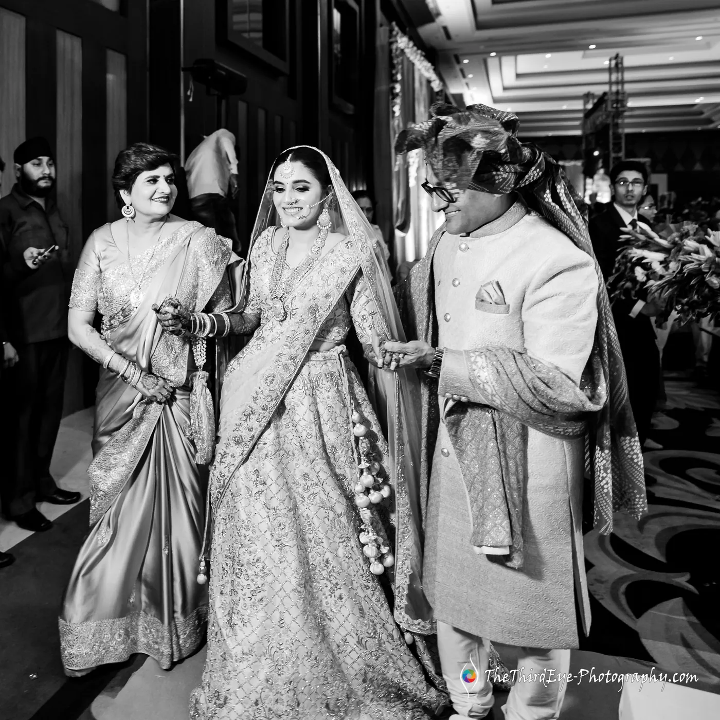 Black and white bride walks forward between a woman in a sari and a man in a light sherwani
