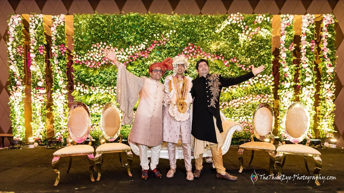 Two men stand gesturing on a green and white floral stage with gold chairs behind them