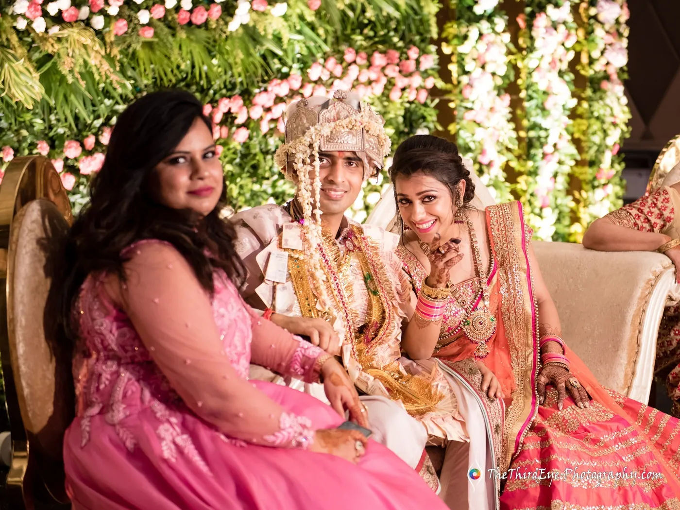 Women in pink and red lehengas sit smiling beside the groom and bride on the floral stage