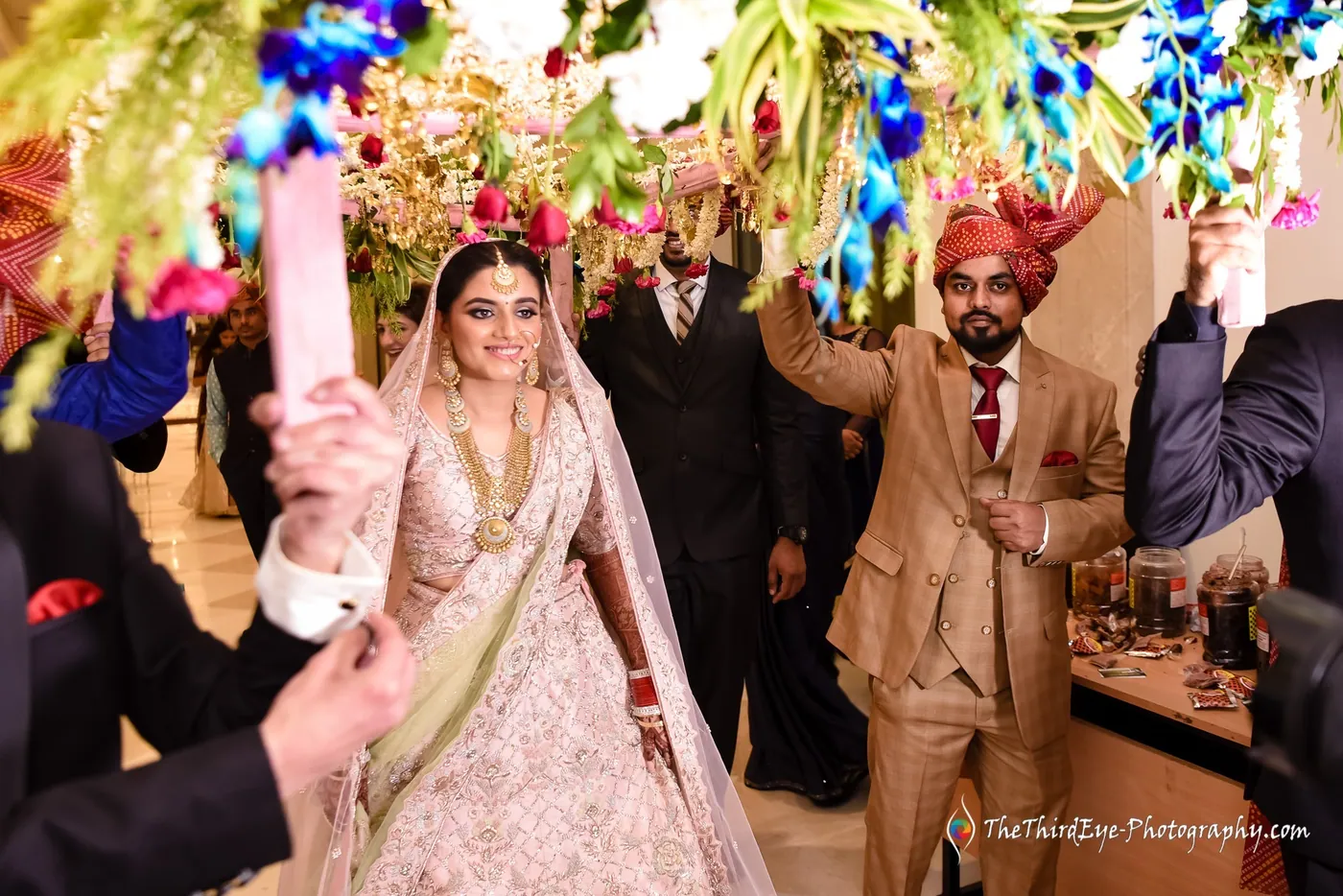 The bride in a pale lehenga walks in as guests film on phones and a man in beige smiles