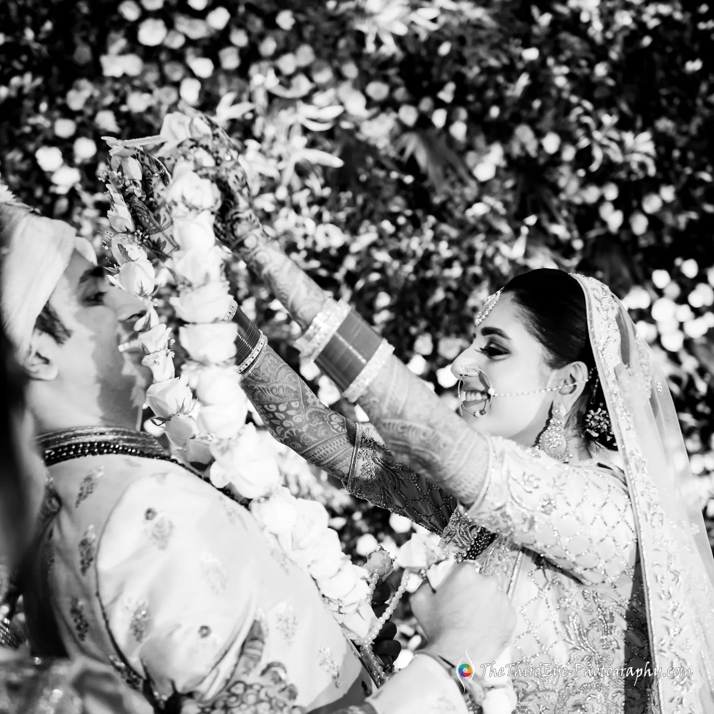 Black and white frame of garlands filling the picture as the bride lifts a mala over the groom