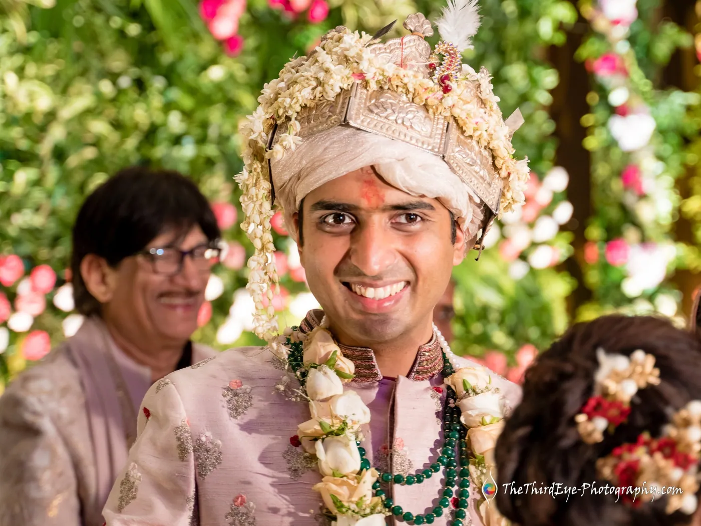 The groom in a flower sehra grins widely with an older man in glasses beside him