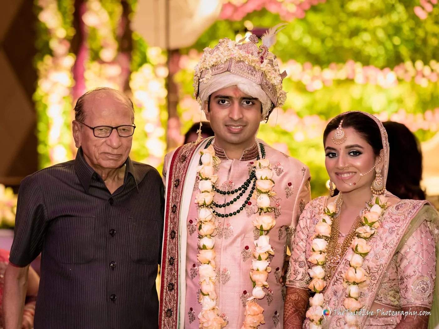The couple stand garlanded with an older man in dark clothes behind them, all smiling toward the camera