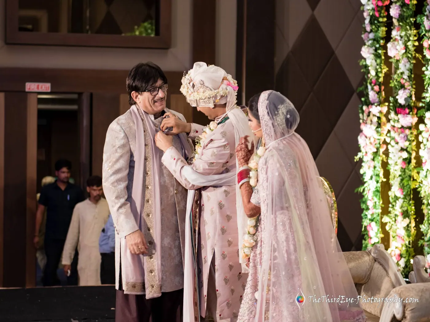 A man pins an ornament on the groom's sherwani as the bride in her veil looks on