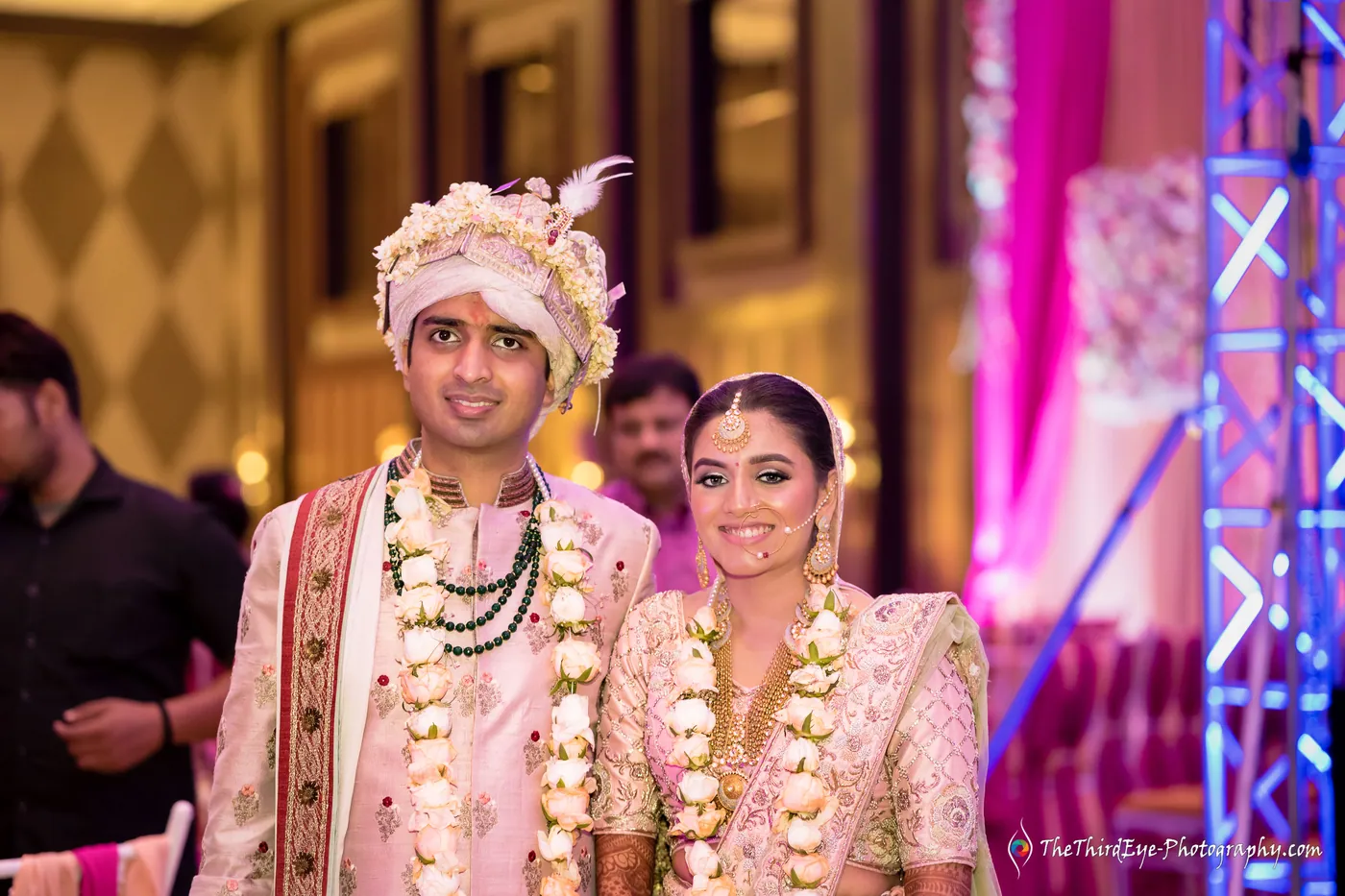 The couple in matching garlands stand smiling in front of pink LED trusses