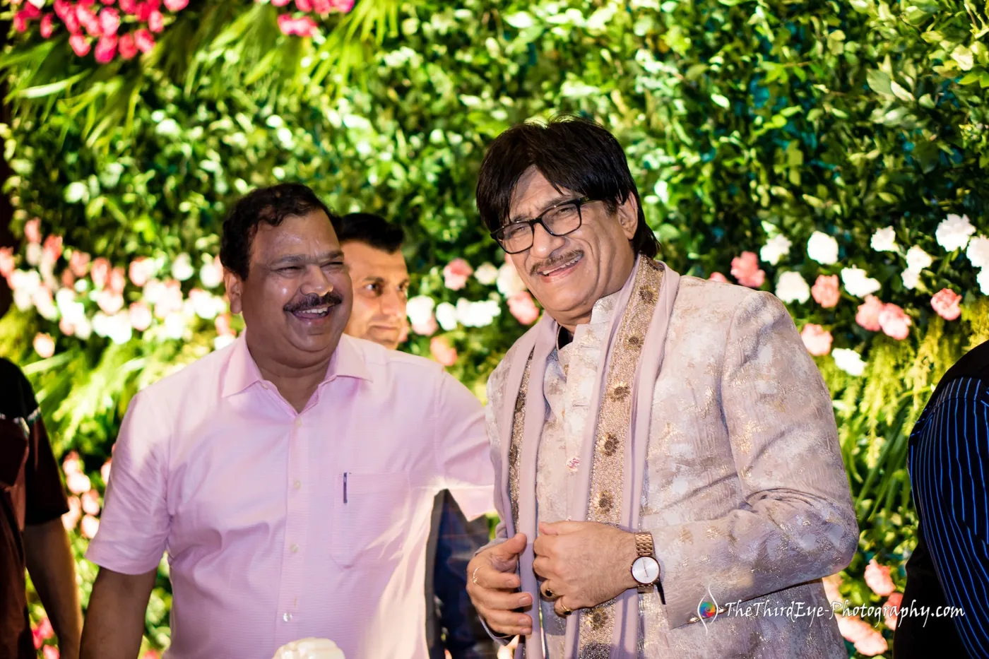 Two men in a floral jacket and a pink shirt smile together on the green floral stage