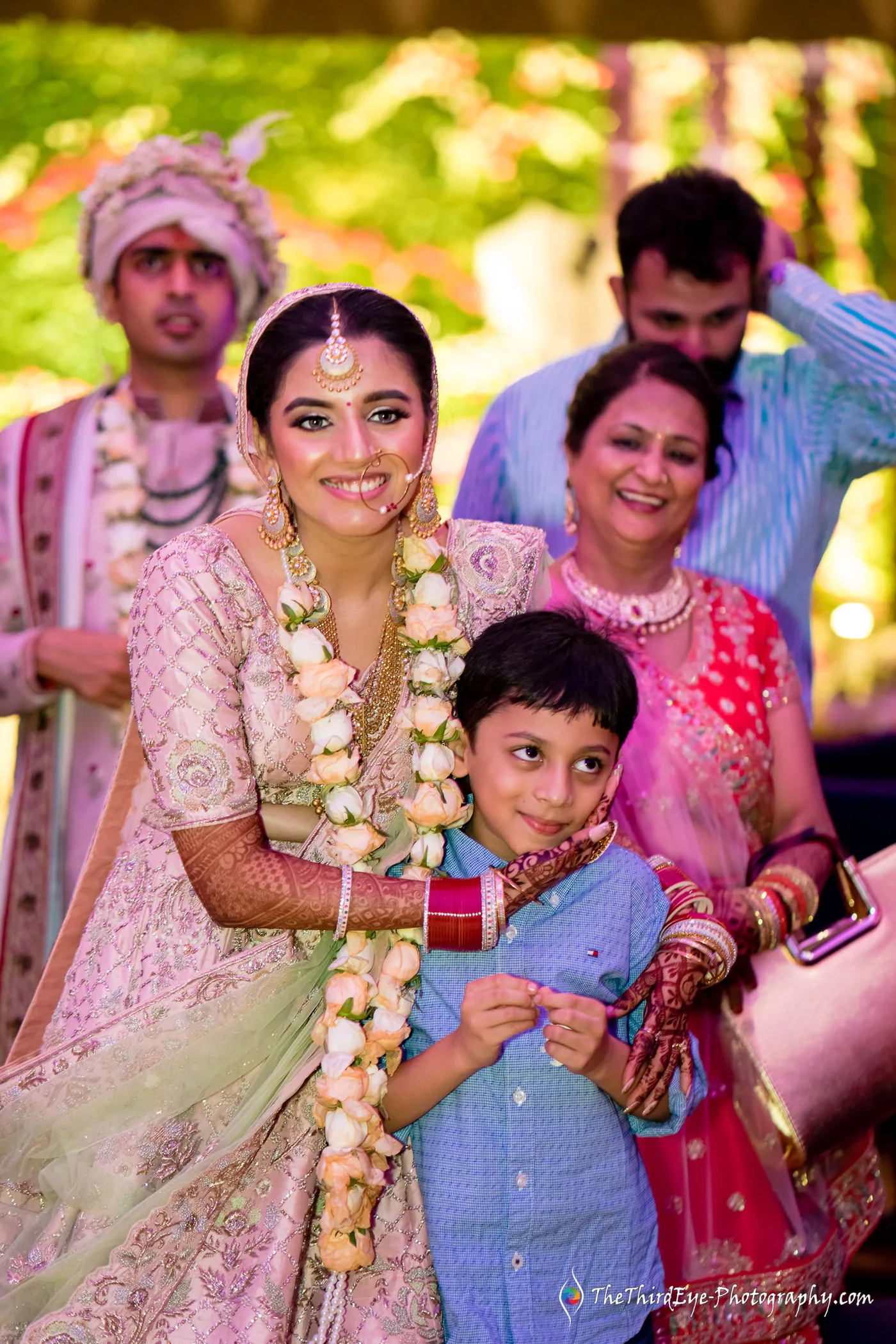 The bride in a pale pink lehenga and garland holds a young boy close as family stand around her