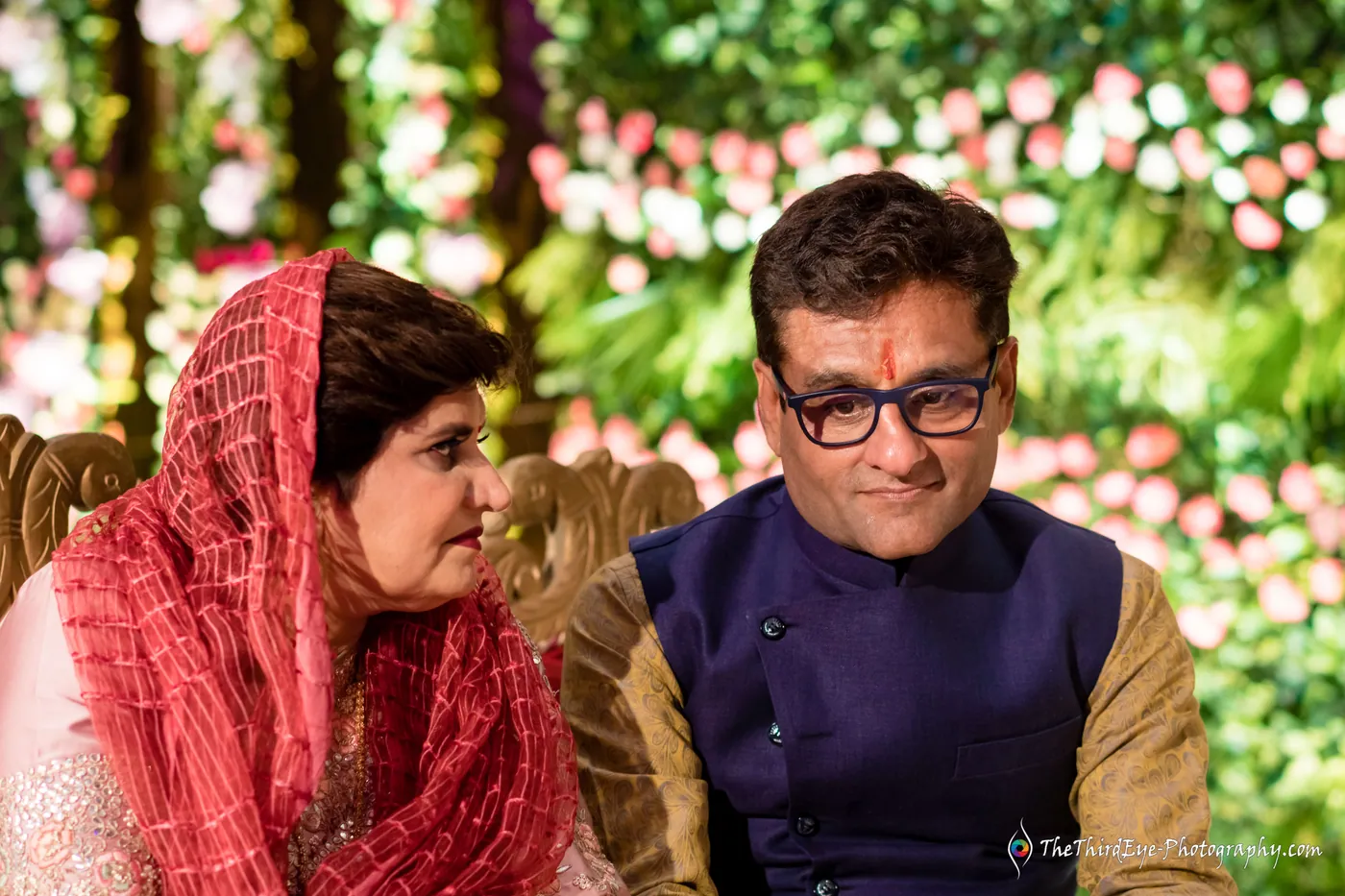A woman in a red dupatta and a man in glasses and a navy waistcoat sit together before flower walls