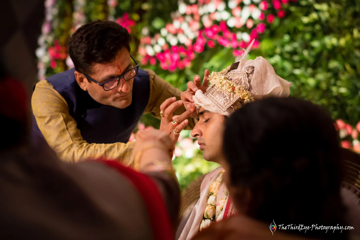 A man in a navy waistcoat applies a tilak to the groom's forehead during the rituals