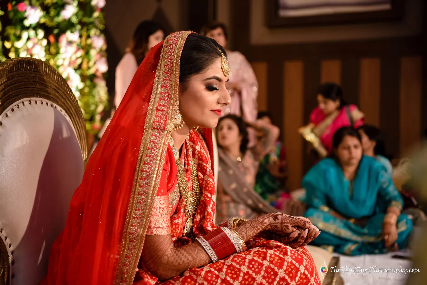 The bride in a red saree sits with downcast eyes during the ritual, relatives seated on the floor behind her