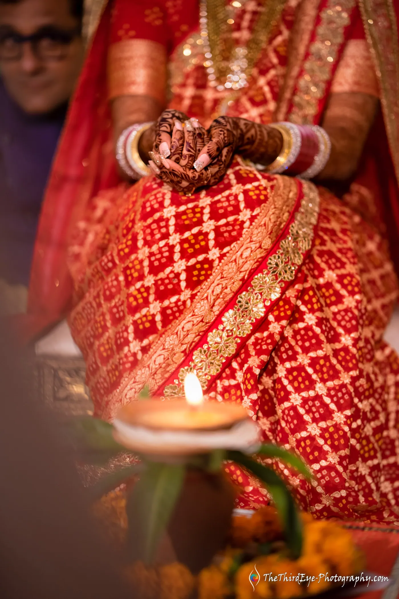 A lit diya sits in the foreground as the bride's henna decorated hands rest on her red patterned saree