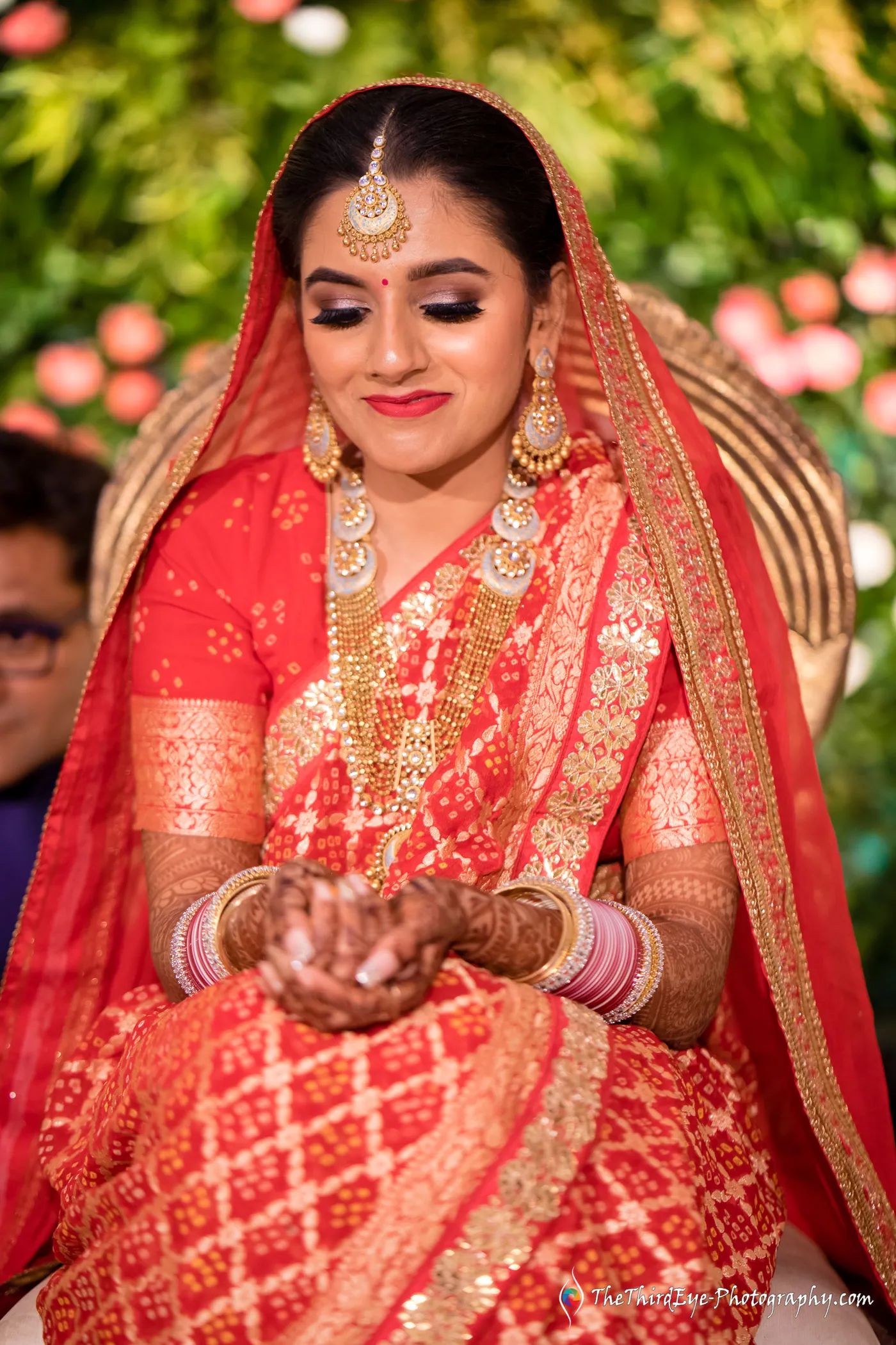 The bride in a red saree and gold jewellery looks down at her palms, smiling gently