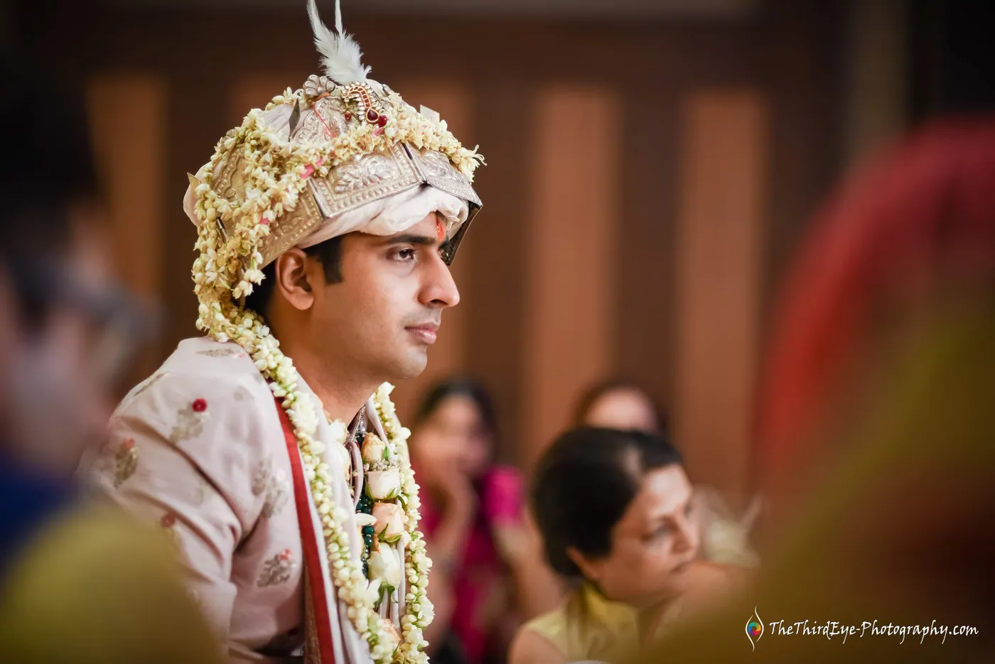 The groom in a white sehra looks off to one side in profile, guests blurred behind him
