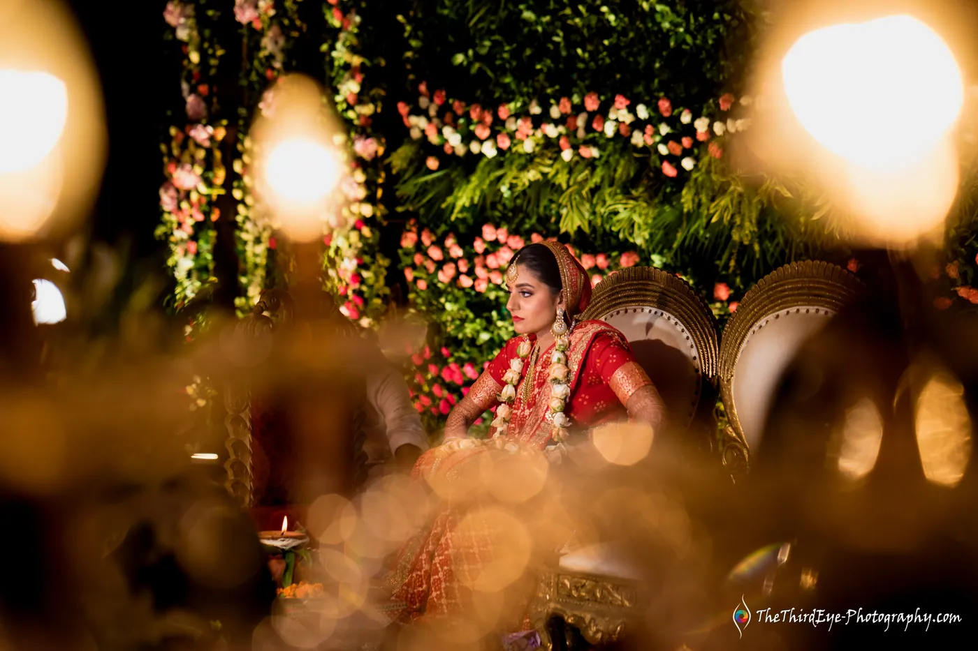 The bride sits on a carved chair in red, glowing in warm light with diyas blurred in the foreground