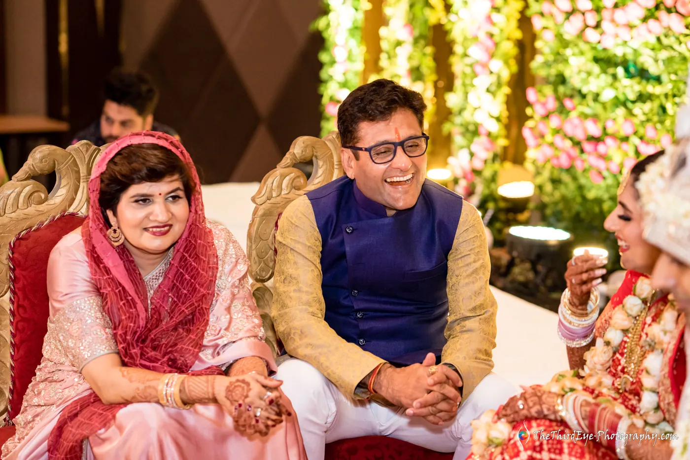 A woman in a pink dupatta and a man in a gold and navy waistcoat sit smiling during rituals