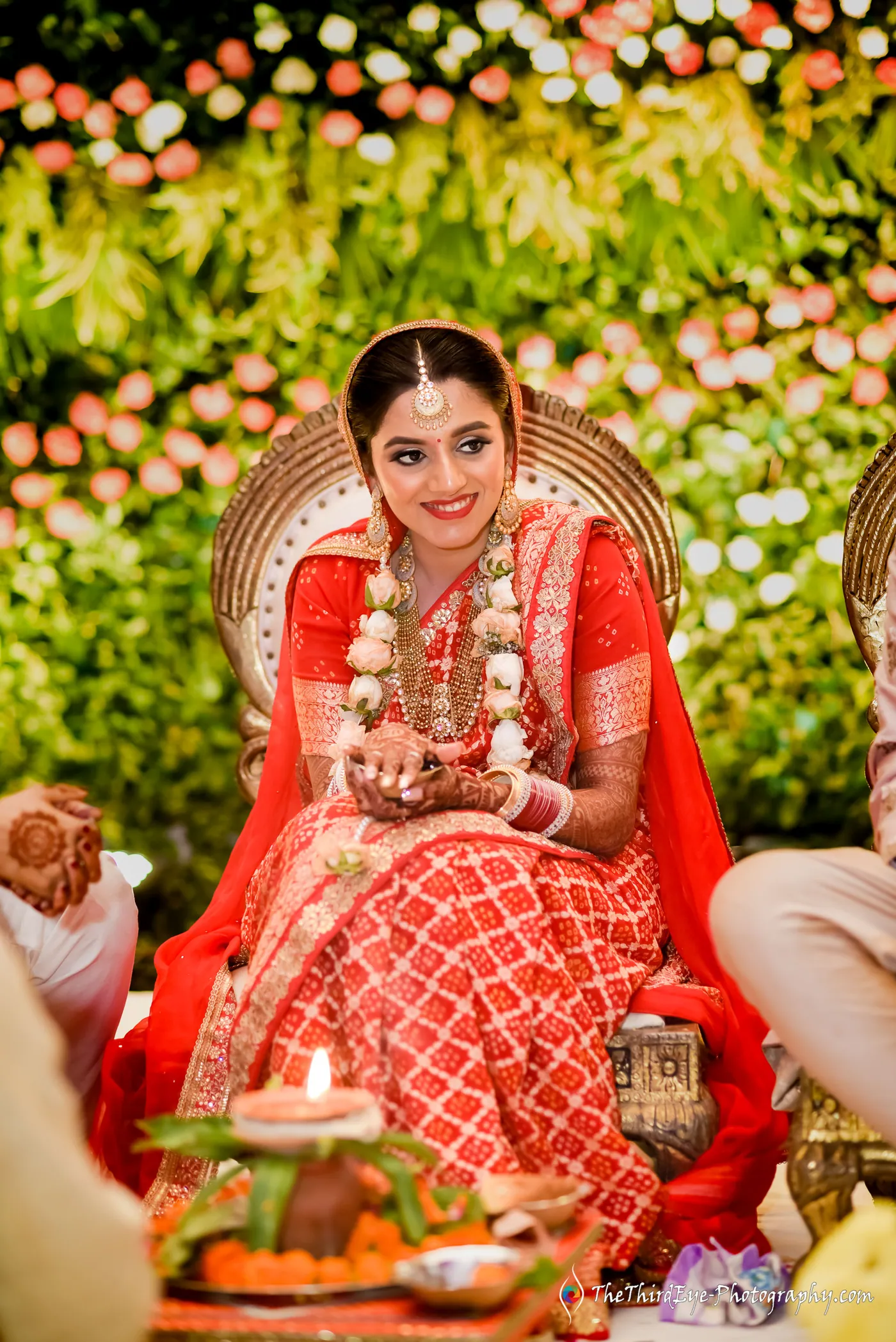 The bride in a red saree sits upright before a flower wall, a diya and puja items at her feet
